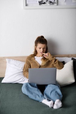 Genç bir kadın modern odasında dinleniyor, bütün gününü aperatif yerken bilgisayarını kullanarak geçiriyor..