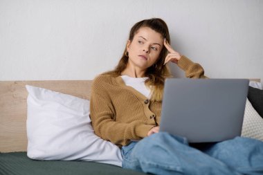 Genç bir kadın, gündelik giysiler içinde yatağında dinleniyor ve düşüncelerini düşünürken dizüstü bilgisayar kullanıyor..