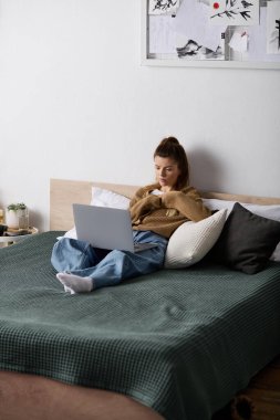 Genç bir kadın dizüstü bilgisayarı ve sıcak bir içecekle rahatça yatakta vakit geçiriyor..