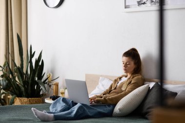 Genç bir kadın, yatağında laptopuyla, huzurlu bir apartman atmosferiyle, serin bir günün tadını çıkarıyor..