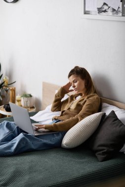 Genç bir kadın gününü rahat bir dairede dizüstü bilgisayarına odaklanmış ve rahatlamış bir şekilde yatakta geçiriyor..