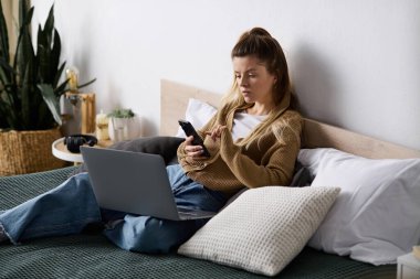 Genç bir kadın yatağında, telefonunu ve laptopunu kullanarak sakin bir günün tadını çıkarıyor..