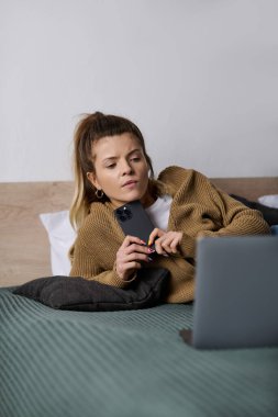 Genç bir kadın şık bir yatak odasında telefonunu ve laptopunu kullanarak yatakta dinleniyor..