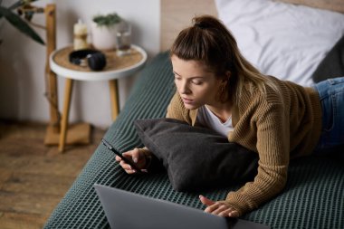 Genç kadın rahat atmosferi kucaklarken telefonunu kullanarak yatakta dinleniyor..