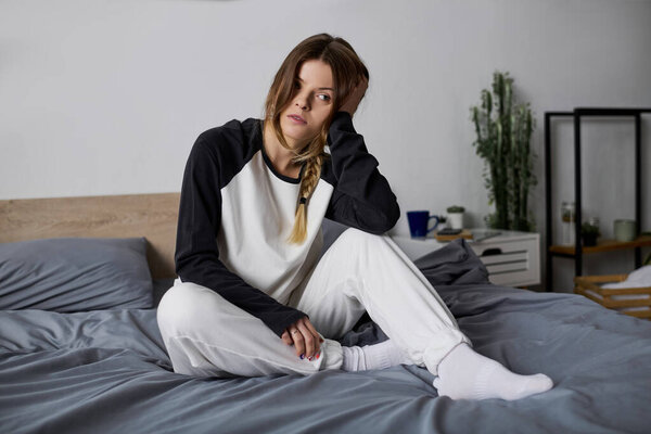 A young woman sits on her bed in a cozy apartment, lost in thought during a restful day indoors.