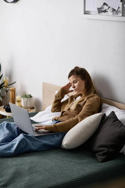 Genç bir kadın gününü rahat bir dairede dizüstü bilgisayarına odaklanmış ve rahatlamış bir şekilde yatakta geçiriyor..