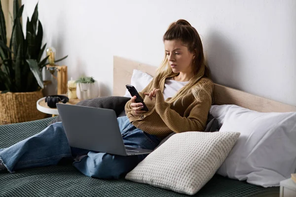 Genç bir kadın yatağında, telefonunu ve laptopunu kullanarak sakin bir günün tadını çıkarıyor..