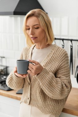 Rahat bir hırka giymiş sarışın bir kadın modern dairesinde sıcak bir içki yudumluyor, rahat hissediyor..