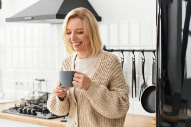 Sarışın bir kadın sıcacık dairesinde sıcak bir içkinin tadını çıkarırken gülümsüyor..