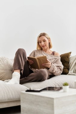 Sarışın kadın rahat elbiseleriyle rahatlar, sıcak ve davetkar dairesinde kitap okur..