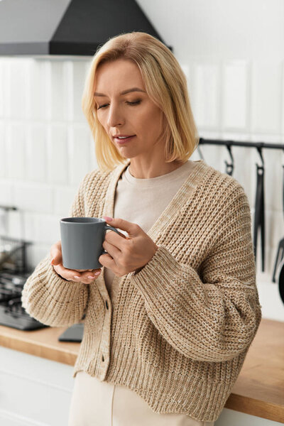 Блондинка в удобном кардигане потягивает теплый напиток в своей современной квартире, чувствуя себя непринужденно.