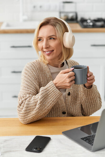 Relaxing in a stylish apartment, a blonde woman sips tea while listening to music in cozy attire.