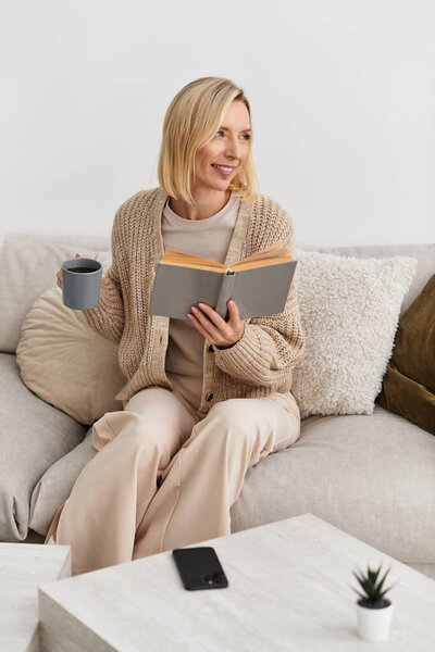 Blonde woman relaxes on a sofa, enjoying a book and warm drink in her cozy, modern apartment.