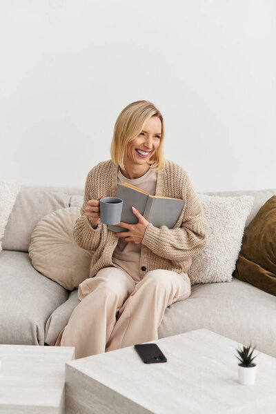 Blonde woman relaxes on a soft couch, reading and sipping tea in a cozy, stylish apartment.