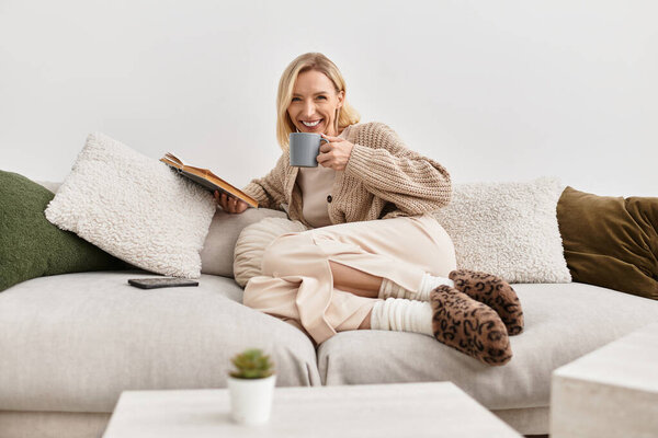 Расслабленная блондинка пьет чай, читая книгу в уютной, современной гостиной.