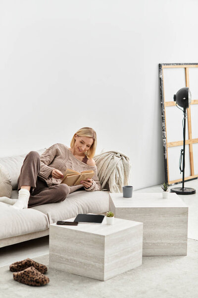 Blonde woman relaxes on a couch, immersed in a book while embracing cozy vibes at home.