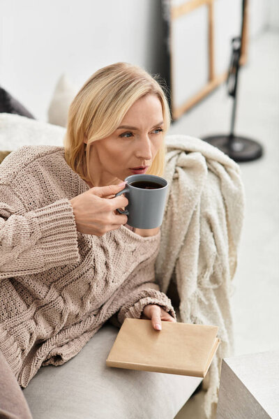 Blonde woman relaxes in her modern apartment, sipping coffee while wrapped in comfort.