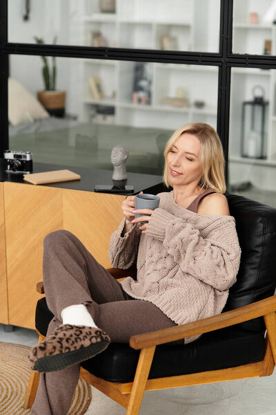 A relaxed blonde woman sits in a comfy chair, sipping tea while dressed in warm, cozy attire.