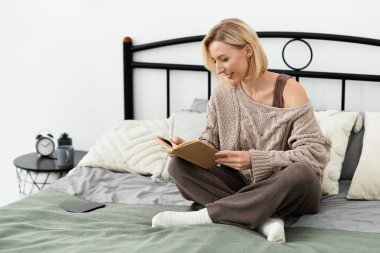 Rahat giyimli bir kadın yatakta rahatlar, evde kitap okur..