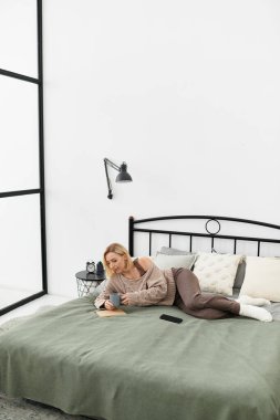 Bir kadın rahat evinde sıcak bir içkinin tadını çıkararak yatağında dinleniyor..