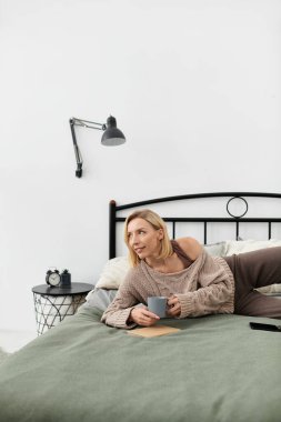 Normal giyinmiş bir kadın yatağına oturur, çayını yudumlar ve huzurlu bir anın tadını çıkarır..