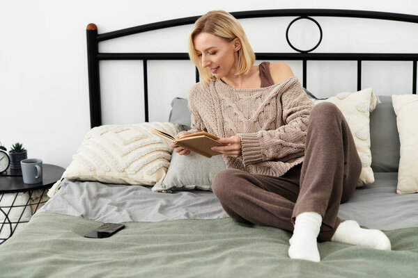 A woman in comfortable clothing relaxes on her bed, reading a book at home.