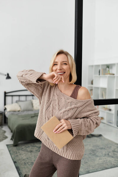 A blonde woman in cozy clothing smiles while spending time at home, embracing relaxation.