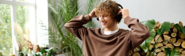 Kıvırcık saçlı genç bir adam, parlak, bitki dolu bir odada müzik dinlerken gülümsüyor..