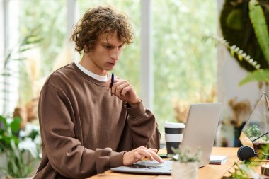 Yeşilliklerle çevrili ahşap bir masada otururken dizüstü bilgisayara odaklanmış genç adam daktilo kullanıyor..