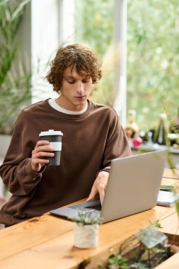 Genç bir adam elinde bir kahve fincanı, parlak yeşil bir alanda dizüstü bilgisayarına odaklanıyor..