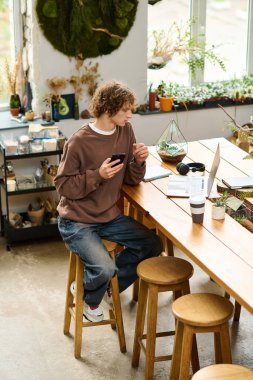 Kıvırcık saçlı genç bir adam telefonunu bitkilerle çevrili canlı bir masada kullanıyor..
