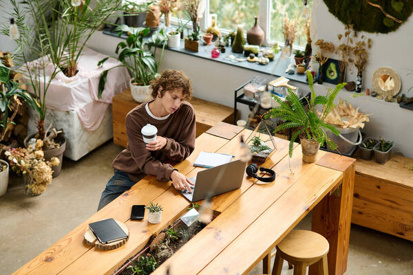 Young man enjoys a cup of coffee while working on his laptop in a plant filled creative space.