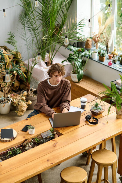Young man focused on his laptop at a wooden table surrounded by greenery in a cozy setting.