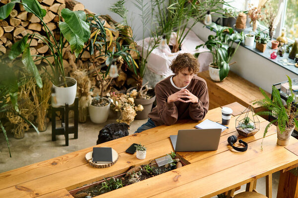 Young man sits at a wooden table, deep in thought, surrounded by greenery and plants.