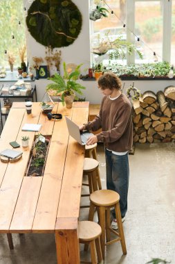 Genç adam yeşilliklerle çevrili şık ahşap bir masada yaratıcı işler yapıyor..