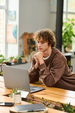 Genç adam dikkatle dizüstü bilgisayarına odaklanır. Parlak bir odada yeşilliklerle çevrilidir..