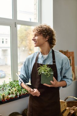 Kıvırcık saçlı genç bir adam kapalı alanda yeşilliklerle çevrili bir saksıyı neşeyle tutuyor..