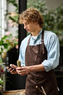 Kahverengi önlüklü genç bir adam yeşillik ve bitkilerle çevriliyken telefonuyla iletişim kuruyor..