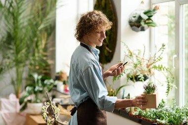 Genç bir adam kapalı alanda canlı bir bahçede bitkilerle ilgilenirken akıllı bir telefonla iletişim kuruyor..