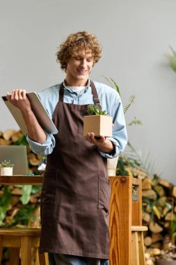 Önlüklü neşeli genç bir adam yeşil, parlak bir alanda gülümserken bir bitki tutuyor..