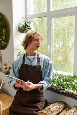 Genç adam, parlak bir kafede elinde tablet tutarken gülümsüyor ve bitkilerle etkileşime giriyor..
