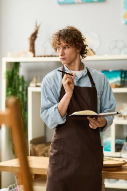 Önlüklü genç bir adam elinde bir defter ve kalem tutarak canlı bir stüdyo ortamında fikir üretiyor..