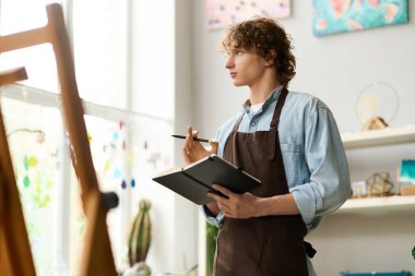 Genç bir sanatçı düşünceli bir şekilde renkli bir stüdyoda duruyor ve defterine fikirler çiziyor..