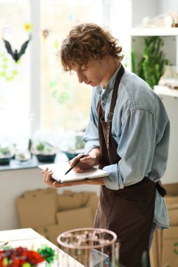 Önlüklü genç bir adam yeşilliklerle çevrili samimi bir sanat stüdyosunda notlar ve eskizler yazıyor..