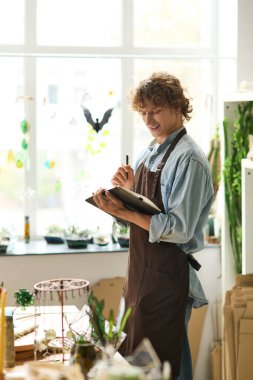 Genç adam sanat malzemeleri ve bitkilerle dolu yaratıcı stüdyosunda bir gülümseme ile tasarımlar çiziyor..