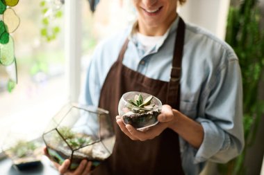 Genç adam elinde bir bitkiyle cam bir akvaryum tutuyor ve sanatsal çalışmalarını gün ışığında gururla sergiliyor..