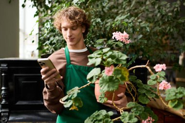 Yeşil önlüklü genç bir adam saksı çiçeği tutuyor ve yemyeşil bir bahçede akıllı telefonunu kontrol ediyor..