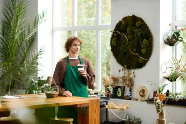 Genç yaratıcı adam, elinde bitki ve dekor dolu bir içkiyle, parlak bir çalışma alanında duruyor..