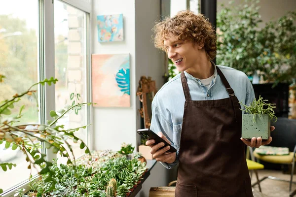 Kıvırcık saçlı genç bir adam, elinde saksı bitkisi ve rahat bir kafede telefon tutarken gülümsüyor..