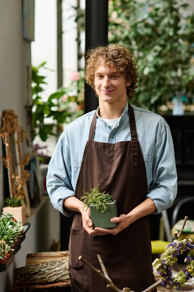 A young man in an apron holds a small plant, smiling amid a lively shop filled with plants.